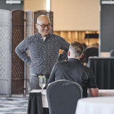 Man stands at table with hands on hips, smiling at a person sitting at a registration table