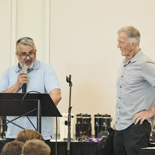 One man stands at podium with a microphone in hand, the other man stand with hand on hip listening